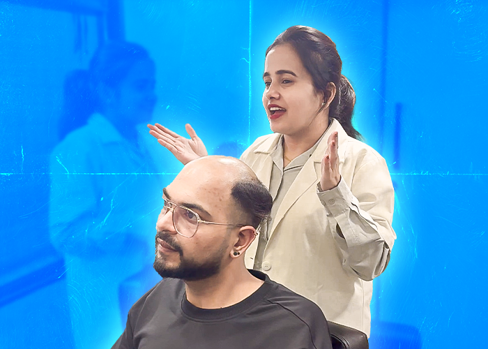 A woman in a beige blazer is giving a consultation to a man with a shaved head, who is sitting in a chair. The woman is gesturing with her hands, appearing to explain or demonstrate something related to the man's hair or scalp. The background is bright blue, with a reflection of the woman visible, suggesting a professional and informative setting. The man is wearing glasses and a black shirt, looking attentive as he listens to the woman.